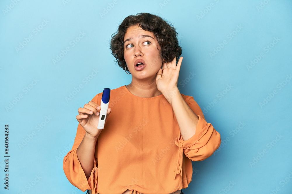 © Asier - Young woman holding pregnancy test, studio background trying to listening a gossip.