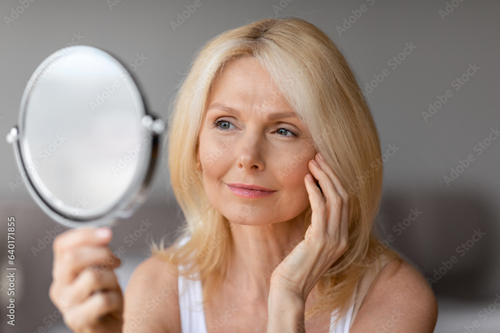 Beautiful middle aged woman touching face and looking in mirror, enjoying her flawless skin, anti-aging beauty routine Beautiful middle aged woman touching face and looking in mirror, enjoying her flawless skin, anti-aging beauty routine