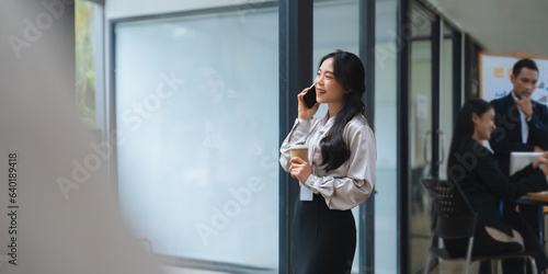 Asian woman are talking on the phone with meeting conference room business people in the background while at her office desk