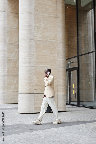 Long shot of young black man in elegant suit moving along buisiness center exterior with huge columns while speaking on smartphone © Seventyfour