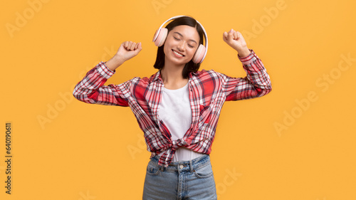 Satisfied asian lady in wireless headphone dancing, enjoying free time, listening music, isolated on yellow background