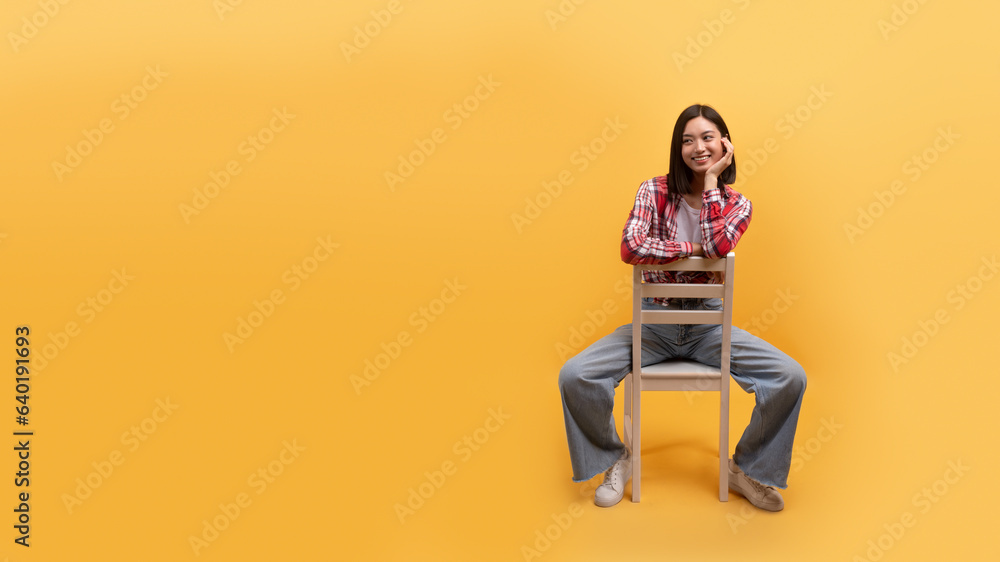 Smiling young chinese lady in casual sitting on chair isolated on yellow background, studio, panorama with free space Smiling young chinese lady in casual sitting on chair isolated on yellow background, studio, panorama with free space