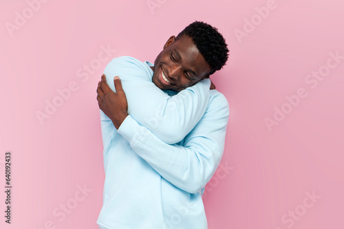 african american man in blue warm sweater hugs and loves himself on pink isolated background photo