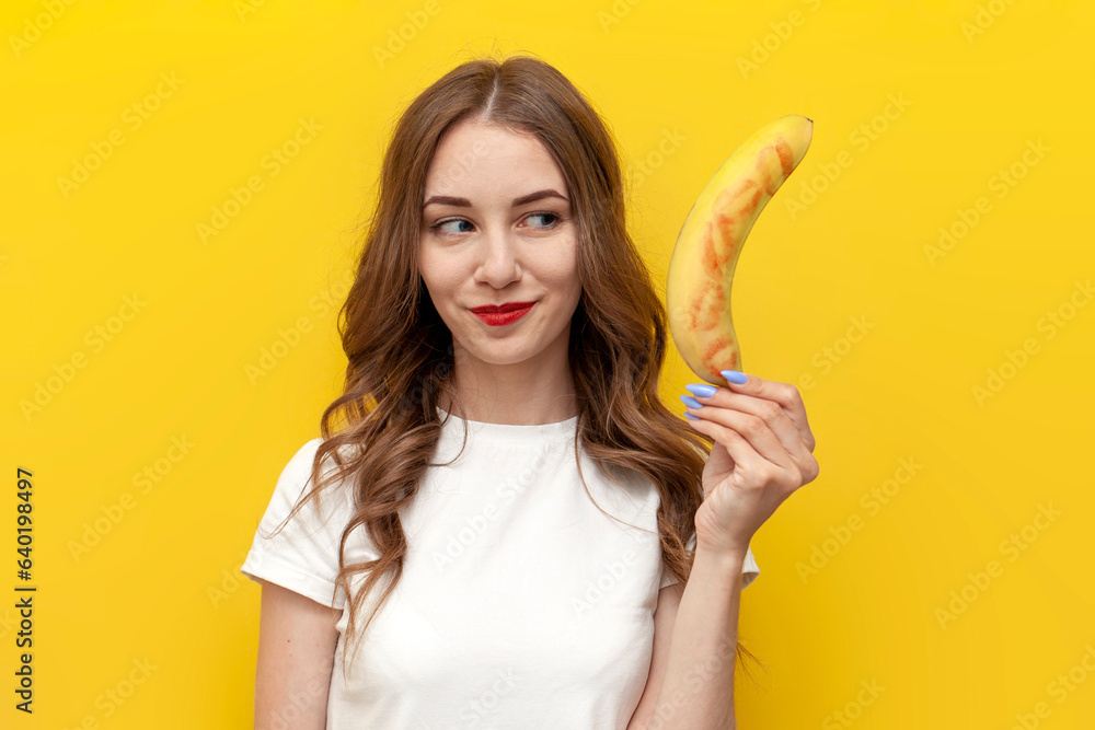 young girl shows kissed banana and hints at intimacy on yellow isolated background, woman with fruit smiles young girl shows kissed banana and hints at intimacy on yellow isolated background, woman with fruit smiles