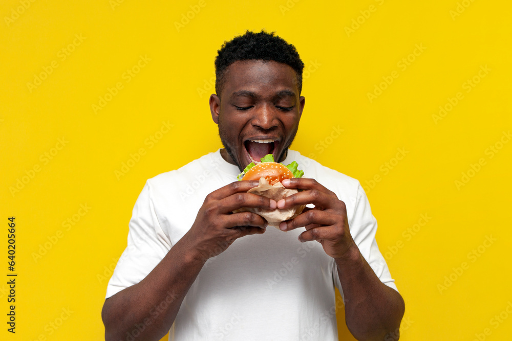 african american man in white t-shirt bites big burger on yellow isolated background, young guy eats fast food african american man in white t-shirt bites big burger on yellow isolated background, young guy eats fast food