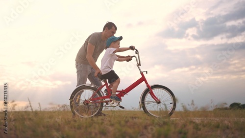 Fototapeta Naklejka Na Ścianę i Meble -  dad teaches son to ride a bike. happy family kid dream concept. the boy sat on bicycle for the first time, his father teaches boy to ride a bicycle. dog runs with lifestyle family, fun family pastime