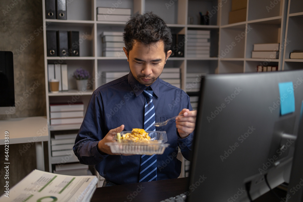 A company employee works overtime after work, he is eating dinner in front of the computer on his office desk in the middle of the night. Overtime and hard work concept.