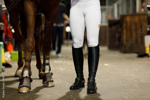 Female legs in black leather boots close up rider jockey walking with horse at stable and preparing horse racing or jumping competition