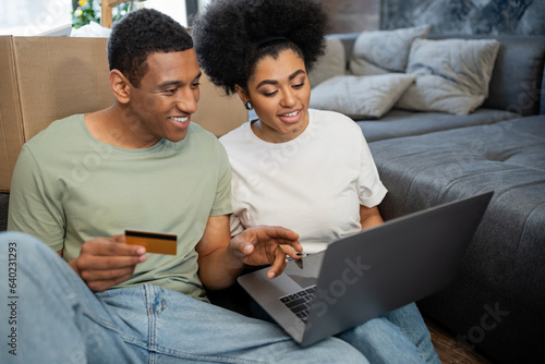 african american couple smiling during online shopping near carton box in living room in new house