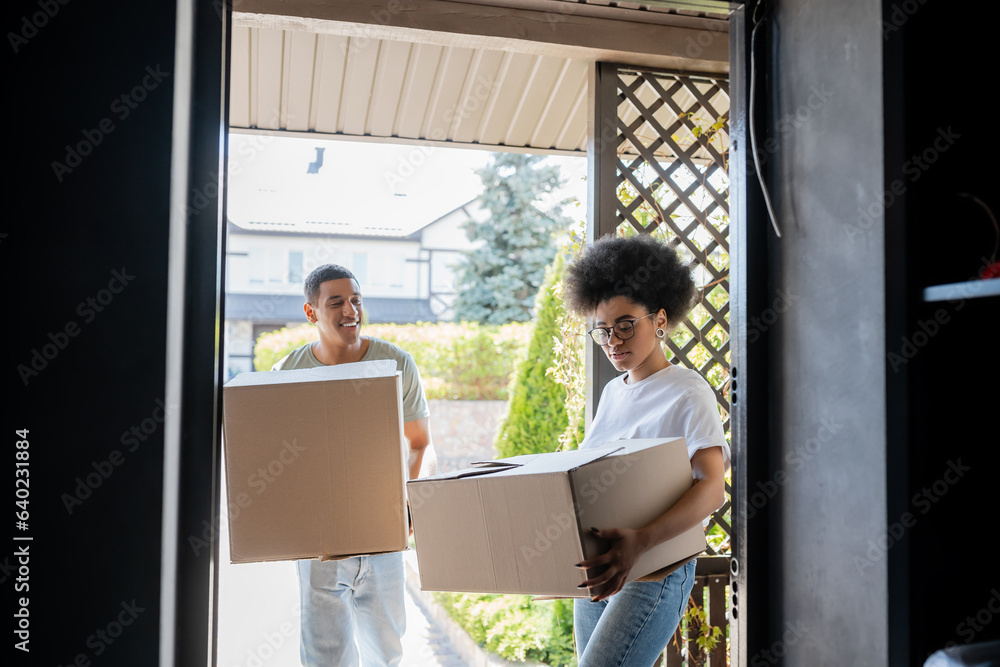 happy african american man holding cardboard box near girlfriend with package and door of new house happy african american man holding cardboard box near girlfriend with package and door of new house