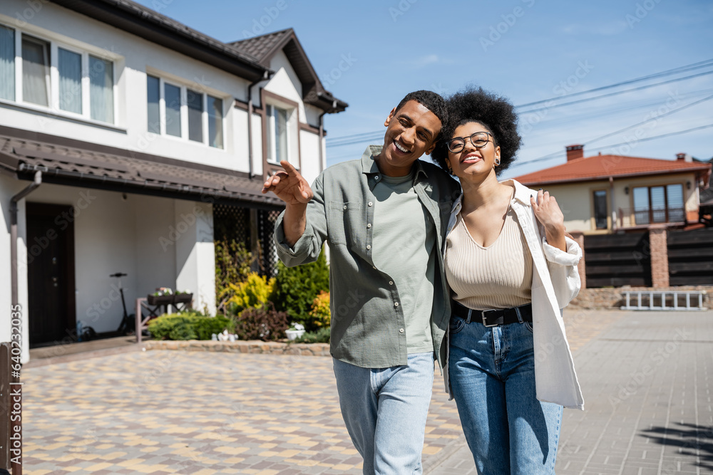 positive african american couple hugging and walking near blurred houses on urban street