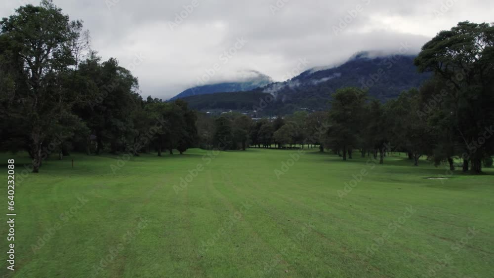 Aerial view of golf field and green forest, mountain under cloudy sky. Drone flying backwards, pull back shot of recreational and relax area for sport and entertainment. Concept of holiday Aerial view of golf field and green forest, mountain under cloudy sky. Drone flying backwards, pull back shot of recreational and relax area for sport and entertainment. Concept of holiday