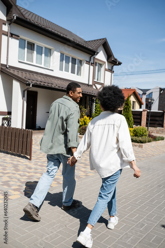 smiling african american couple holding hands while walking on sidewalk near houses on street
