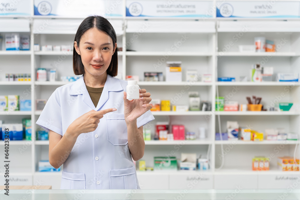 Happy beautiful asian female pharmacist wearing lab coat standing and pointing finger to pill bottle, She looking at camera feels good, trustworthy and proud of his work in the pharmacy drugstore. Happy beautiful asian female pharmacist wearing lab coat standing and pointing finger to pill bottle, She looking at camera feels good, trustworthy and proud of his work in the pharmacy drugstore.
