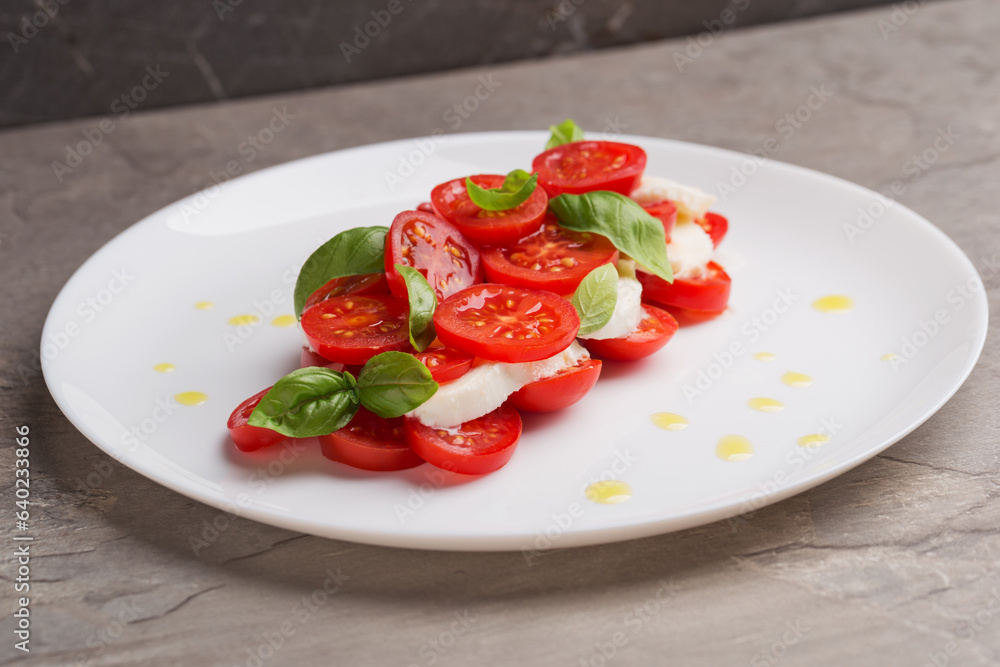 A view of caprese salad with tomatoes, mozzarella, basil and olive oil on white plate on dark stone table. Isometric style. Mediterranean cuisine. Selective focus. A view of caprese salad with tomatoes, mozzarella, basil and olive oil on white plate on dark stone table. Isometric style. Mediterranean cuisine. Selective focus.