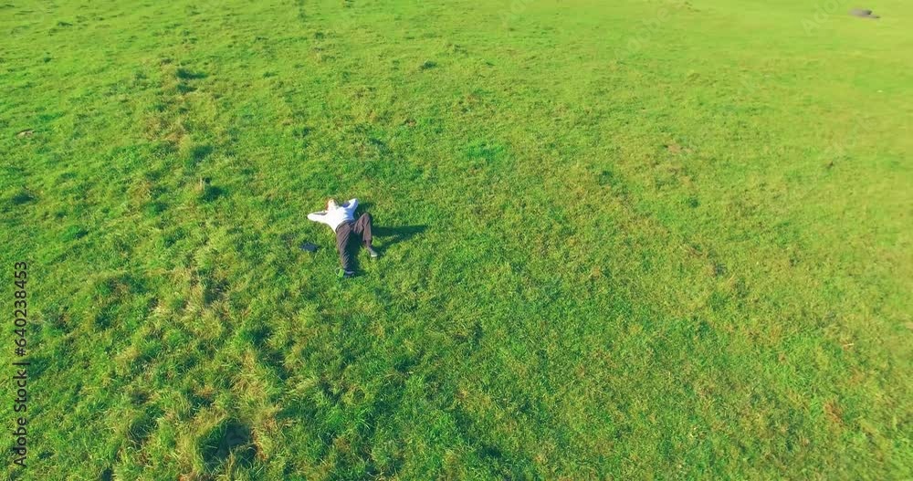 Aerial view point. Low orbital flight around man lied on green grass with notebook (pad) at yellow rural field. Casual weared. Sun rays. Radial movement.