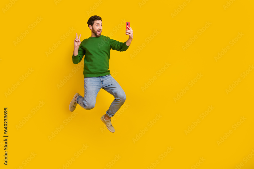 Full body photo of video selfie shooting popular blogger show v sign gesture hold smartphone jumping isolated on yellow color background