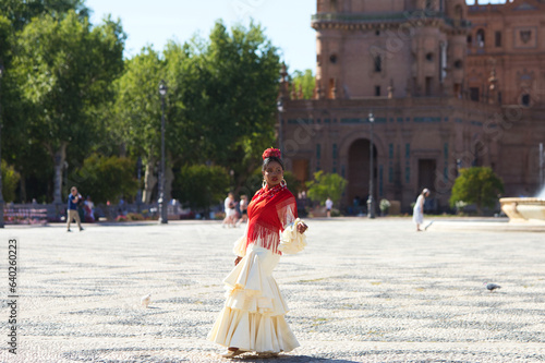 Young black woman dressed as a flamenco gypsy in a square in Seville, Spain. She wears a beige dress with ruffles and a red shawl with flowers. Flamenco cultural heritage of humanity. Dance concept.
