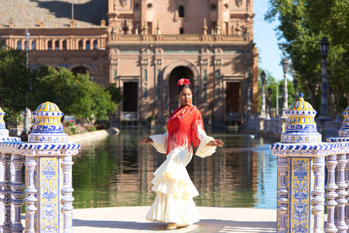 Young black woman dressed as a flamenco gypsy in a famous square in Seville, Spain. She is wearing a beige dress with ruffles and a red shawl and is in front of a canal in the square. © @skuder_photographer