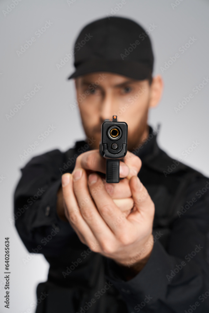 Unrecognizable cop in uniform taking aim, focused on camera indoors. Close up of black weapon, gun, held by blurred police officer silhouette, on gray background. Concept of danger, weapon, target. Unrecognizable cop in uniform taking aim, focused on camera indoors. Close up of black weapon, gun, held by blurred police officer silhouette, on gray background. Concept of danger, weapon, target.