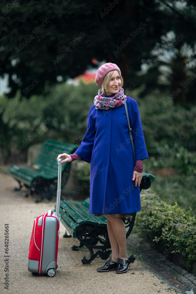 There is a woman walking down the street with a red suitcase in her hand.
