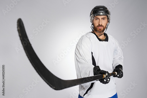 Bearded hockey player in protective shield pointing black stick into camera. Close up view of caucasian male ice hockey sportsman in uniform performing slap shot, isolated on gray. Concept of sport. photo
