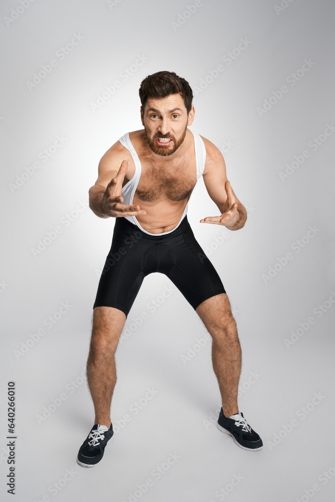 Obraz premium Young wrestler in black and white tight singlet getting ready to attack in studio. Front view of bearded athlete making face, while staying in wrestling stance, isolated on white. Concept of sport.