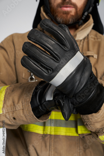 Unrecognizable bearded firefighter wearing black protective gloves. Crop view of male fireman putting on leather firefighting gloves, on white background. Concept of profession, fire equipment.