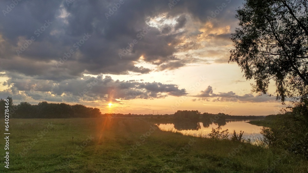 Summer evening, the sun is sinking below the horizon. Colorful cloudy sky and trees are reflected in the quiet river water. A feeder rod is set up on the grassy bank to catch fish. A forest grows