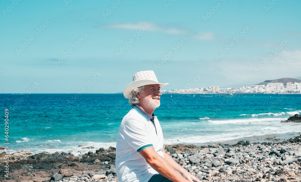 Happy white-haired senior man enjoying sea vacation sitting at the beach in a sunny day