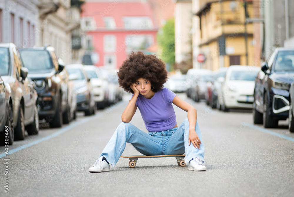 Young ethnic Moroccan woman sitting on skateboard on street near parked cars