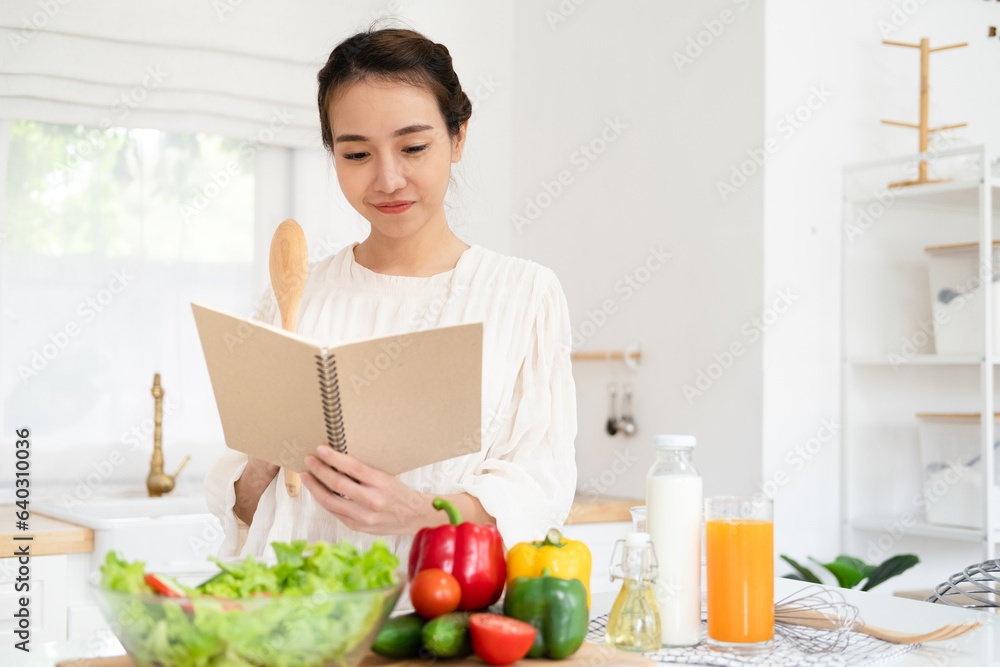 Smiling housewife woman looking at recipe in cookery book preparing vegetable salad cooking food in light kitchen at home. Dieting healthy lifestyle concept.