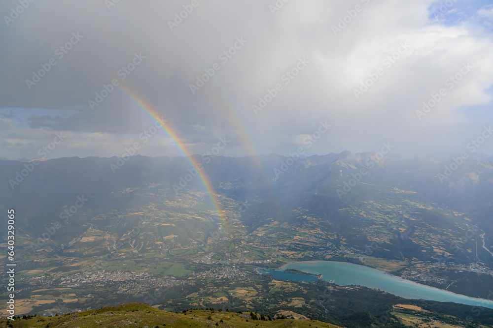 A scenics view of a mountain summit with a majestic double rainbow and Embrun, Hautes-alpes, France in the background under a stormy and rainy weather conditions A scenics view of a mountain summit with a majestic double rainbow and Embrun, Hautes-alpes, France in the background under a stormy and rainy weather conditions