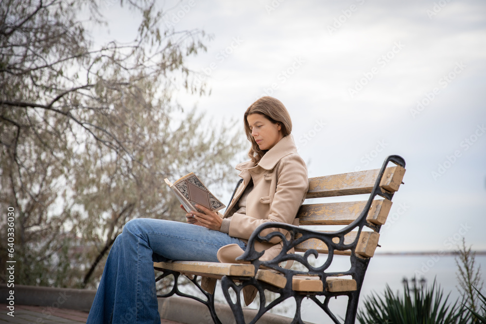 Young beautiful slim woman in a light coat and blue jeans is reading a book on a bench in the city.