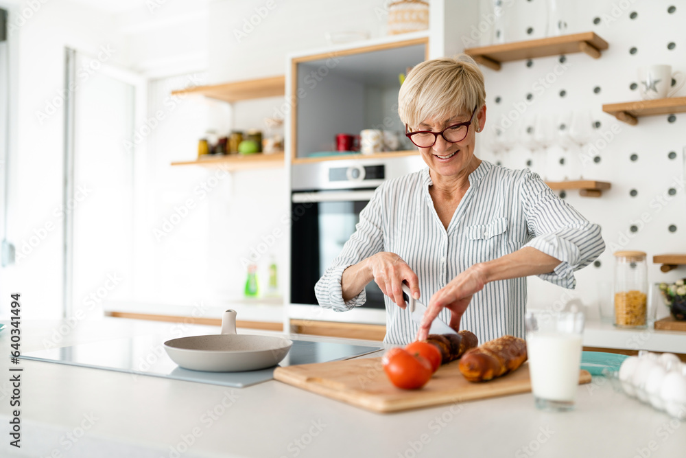 Joyful elderly blonde woman cutting vegetables on kitchen board while drinking milk in her kitchen.