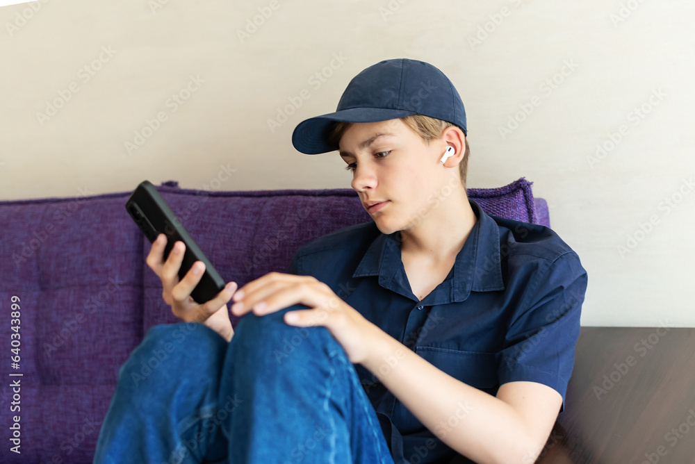 Teenager boy relax sit on sofa at home using cellphone - texting chatting with friend, listening music with earphones and mobile phone, shop online or check mobile application or social media
