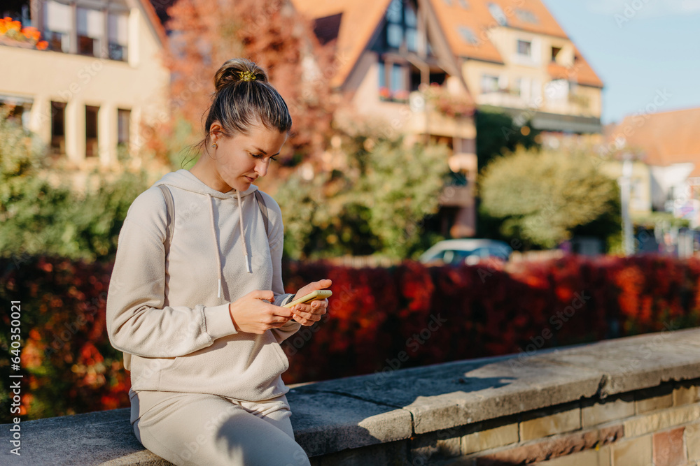 Obraz premium Young Fashionable Teenage Girl With Smartphone In Europian Park In Autumn Sitting At Smiling. Trendy Young Woman In Fall In Park Texting. Retouched, Vibrant Colors. Beautiful Blonde Teenage Girl