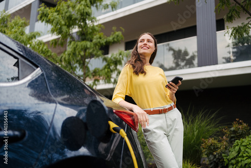 Close up of beautiful woman using smartrphone while charging her electric car on the street. photo