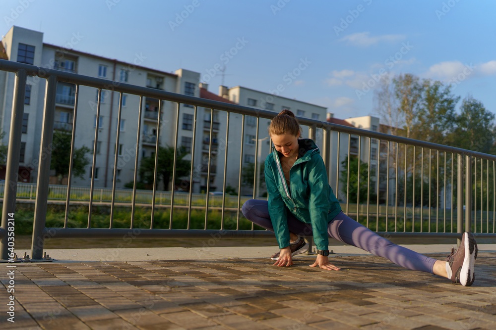 Attractive young woman stretching before her early morning run along the embankment of river.