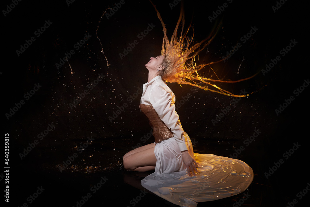 Fototapeta premium Beautiful sexy skinny girl and her flying hair in a dark hall with splashes of water. Female Model blonde woman posing with water on a black background in the studio