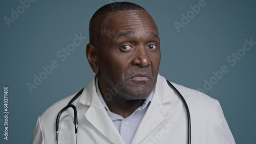 Close-up unsure confused ethnic african american man doctor surgeon in medical white coat in blue background studio shrug shake head making I don't know gesture no answer waving negative disapproval