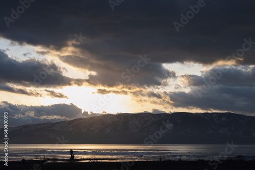 Fototapeta Naklejka Na Ścianę i Meble -  a couple stands at the edge of a lake with mountains in the background at sunset