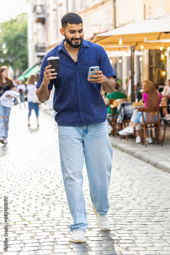 Happy indian man use smartphone typing text messages browsing internet social media web app working chatting online enjoying morning to-go coffee hot drink outdoor. Guy tourist walking in city street © Andrii Iemelianenko