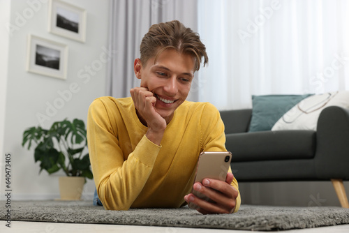 Happy young man having video chat via smartphone on carpet indoors