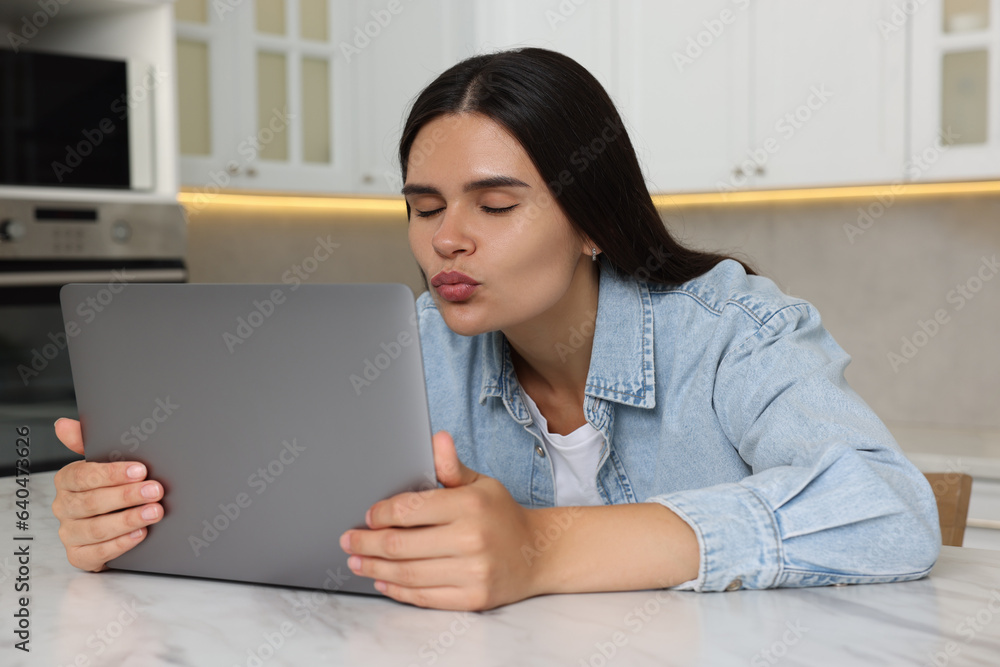 Young woman having video chat via laptop and blowing kiss at table in kitchen Young woman having video chat via laptop and blowing kiss at table in kitchen