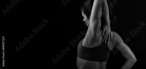 Female sporty muscular woman doing stretching relaxing workout of the shoulders, blades in sport bra, holding the neck the hand on black studio background with empty copy space. Back view. Black