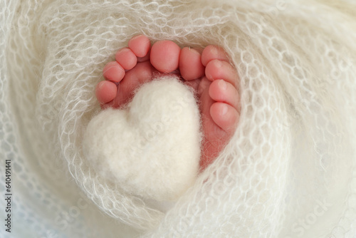 Knitted white heart in the legs of a baby. The tiny foot of a newborn baby. Soft feet of a new born in a white wool blanket. Close up of toes, heels and feet of a newborn. Macro photography © Vad-Len