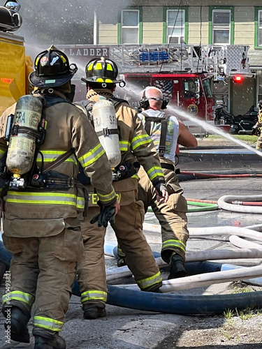 Emergency situation pictured. Firemen in full gear with oxygen tanks are standing in a street. The firefighters are spraying water putting out a house fire. Firetrucks can be seen surrounding the men.