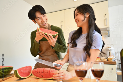 Happy married couple in aprons standing behind wooden kitchen tabletop and eating sweet watermelon
