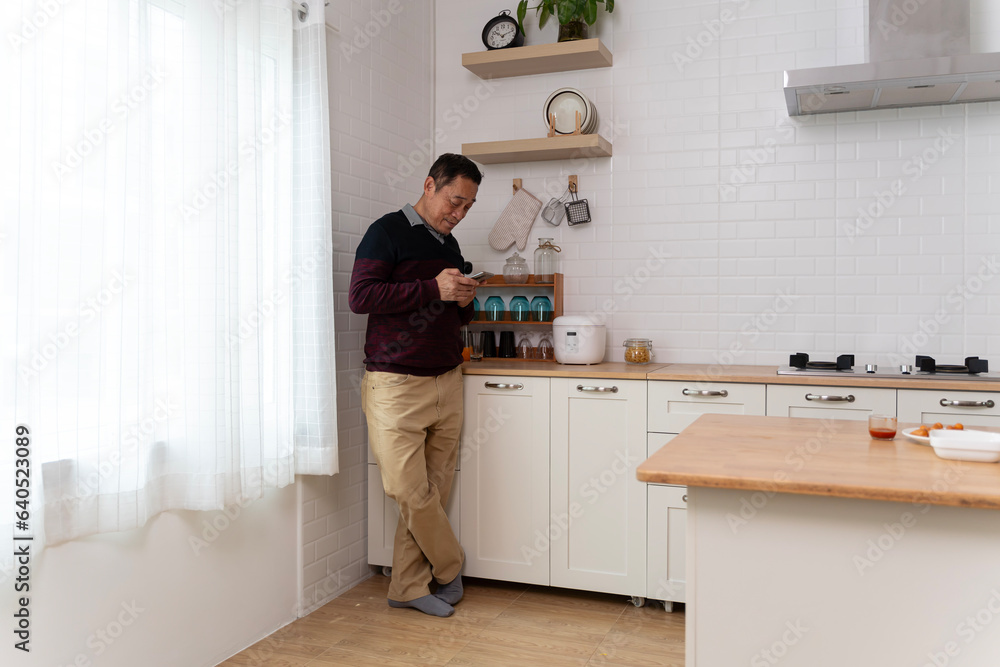 Happy middle aged old retired man in eyewear using cellphone, checking recipe online for tasty food, setting timer preparing meal alone in modern kitchen. Happy middle aged old retired man in eyewear using cellphone, checking recipe online for tasty food, setting timer preparing meal alone in modern kitchen.